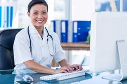 Happy doctor using her computer and looking at camera in medical office