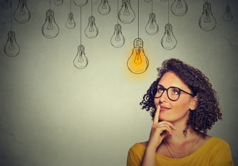 Thinking woman in glasses looking up with light idea bulb above head isolated on gray wall background